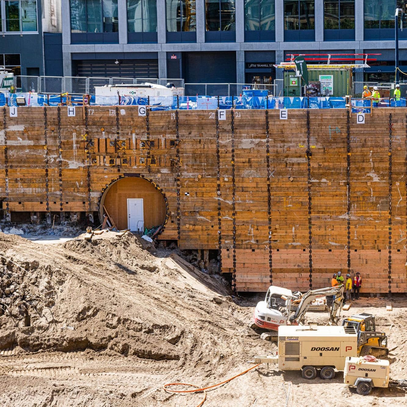 A large shoring wall on the press blocks construction site features a man door for tunnel access to the adjoining building